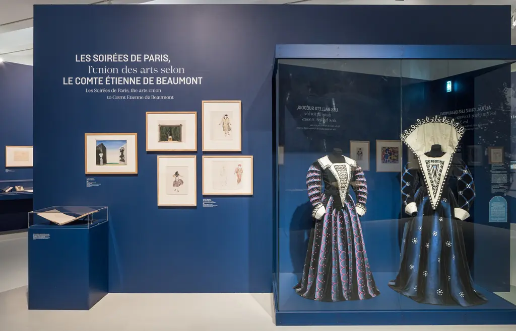 Sur un mur bleu roi, le texte "Les soirées de Paris, l'union des arts selon le comte Étienne de Beaumont", plusieurs dessins et devant, deux vitrines montrant un livre ouvert et deux robes de théâtre.