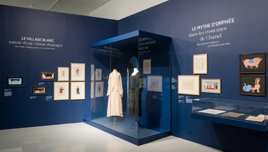 Sur des murs bleu roi, deux textes : "Le village blanc, miroir d'une vision d'époque" et "Le mythe d'Orphée dans les costumes de Chanel". Sur les murs, des dessins et des maquettes de scénographie de théâtre. Devant, une vitrine montrant deux costumes de théâtre.