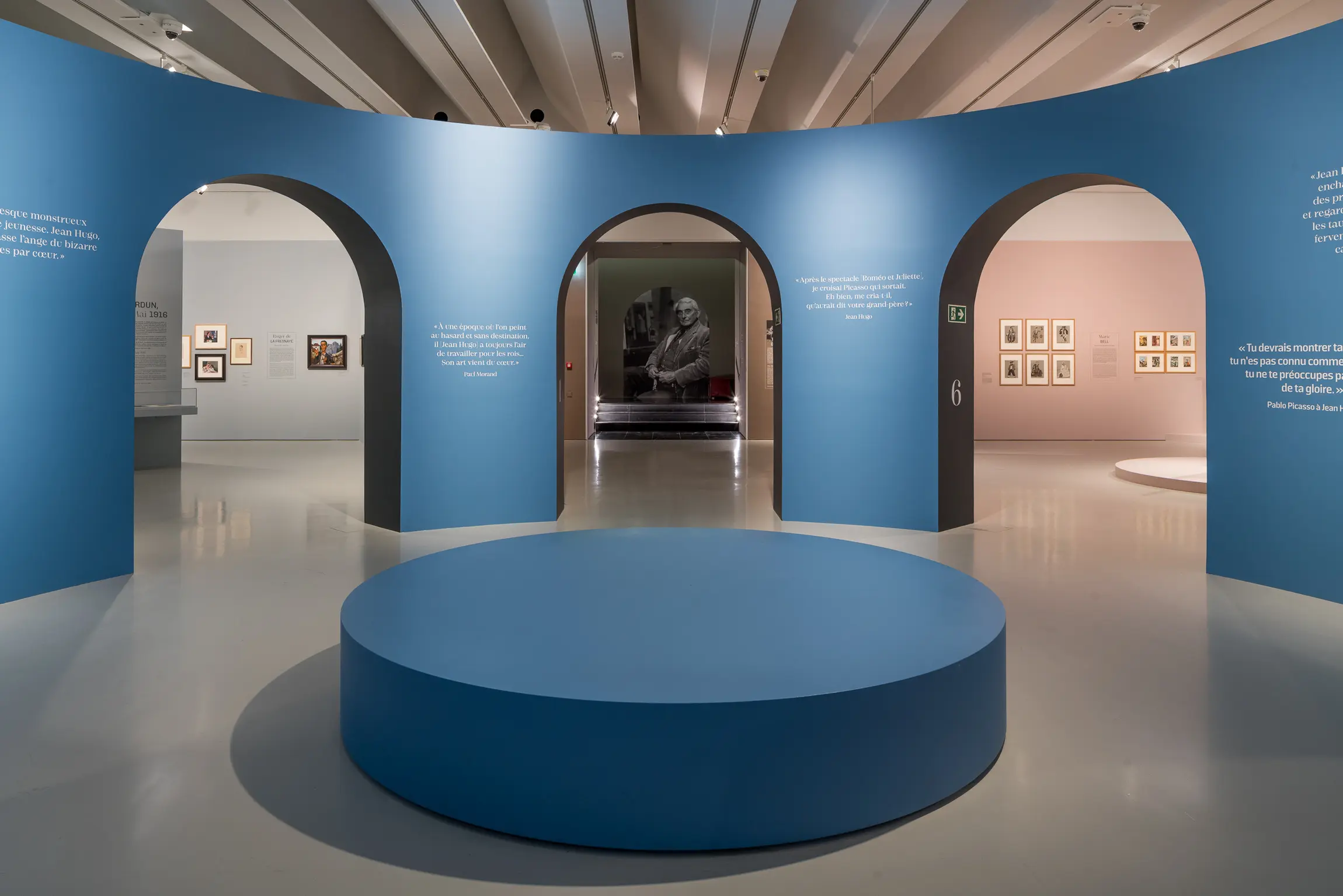 Dans l'espace central de l'exposition. Au premier plan, un grand banc bleu circulaire. Derrière, un mur arrondi bleu avec trois arches menant vers d'autres salles. En fond de l'arche centralee, on aperçoit une photo de Jean Hugo âgé.