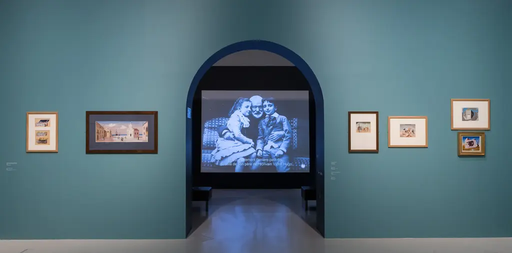 Un mur bleu clair sur lequel sont accrochées plusieurs œuvres de Jean Hugo. Au centre, une arche laisse apparaître une autre salle dans laquelle est projetée une vidéo.