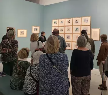 Un groupe de visiteurs écoute une visite guidée dans une salle de l'exposition Jean Hugo. Les murs sont bleu clair.