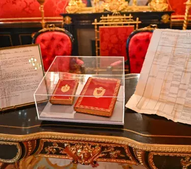 Sur un bureau en bois aux moulures dorés, quatre documents sont présentés : à gauche, une attribution de titre et de rente rédigée et signée par Cambacérés ; à droite, un arrêté du comité de salut public de 1794 ; au centre, sous cloche, deux volumes de l'exemplaire de Napoléon de la conférence du Code Civil 1804.