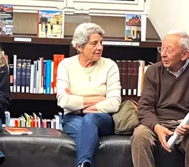 Dans une bibliothèque, devant une étagère remplie de livres, deux femmes et un homme discutent sur un banc.