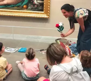 Dans une salle du musée, une médiatrice tend un bouquet de roses (rouges et blanches) à une petite fille qui fait partie d'un groupe d'enfants assis.